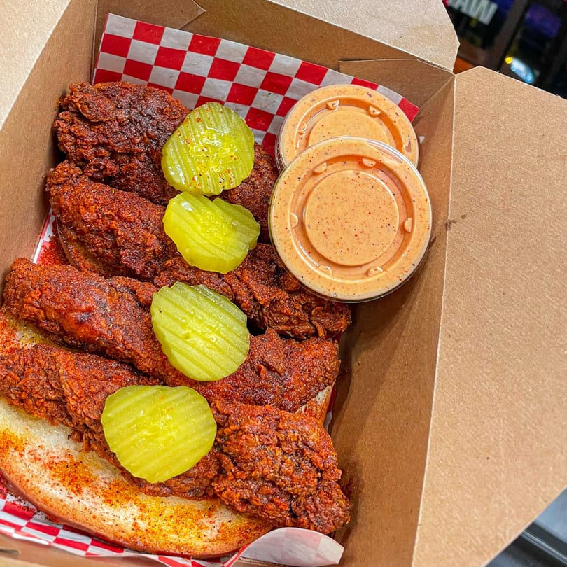 Nashville Coop's hot fried chicken tenders with pickles and dipping sauce in a takeout box.
