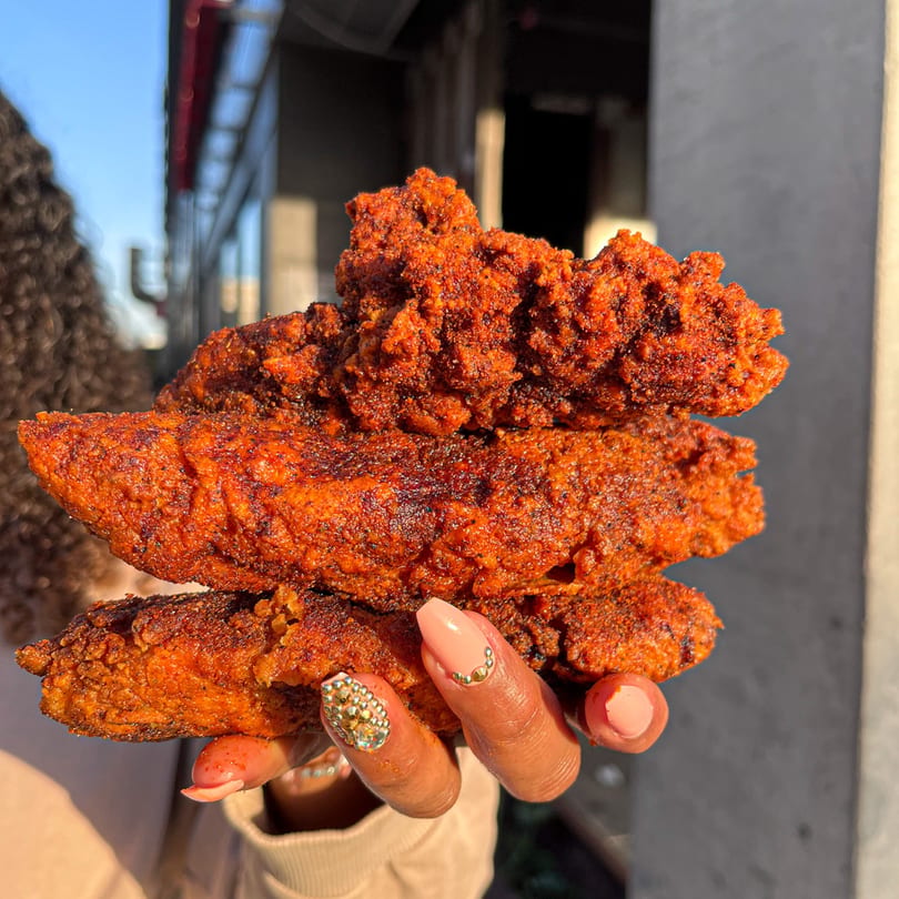 A hand holds up three crispy, red Nashville Coop fried chicken tenders against a sunny outdoor background.