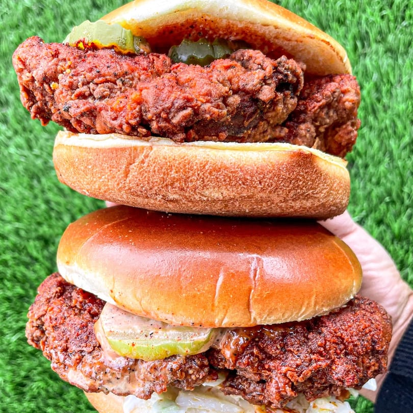 Two Nashville Coop fried chicken sandwiches stacked, with pickles, sauce, and coleslaw, against green.