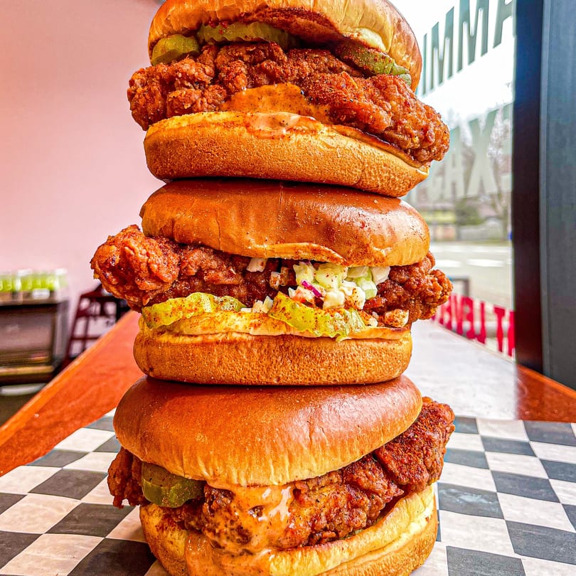 A towering stack of three Nashville Coop fried chicken sandwiches with pickles and sauce.