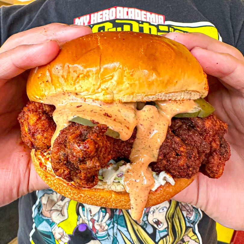 Hands holding a Nashville Coop fried chicken sandwich with sauce, pickles, and slaw.